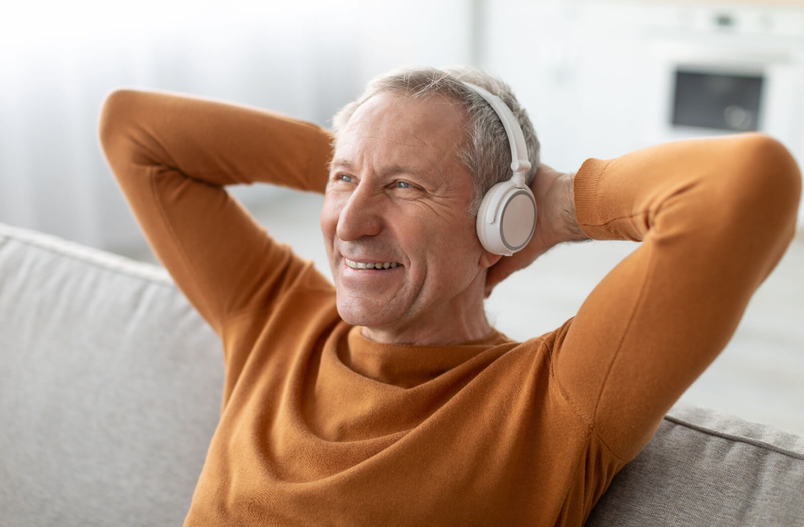 A grinning older adult relaxing with their hands behind their head while listening to an audiobook.