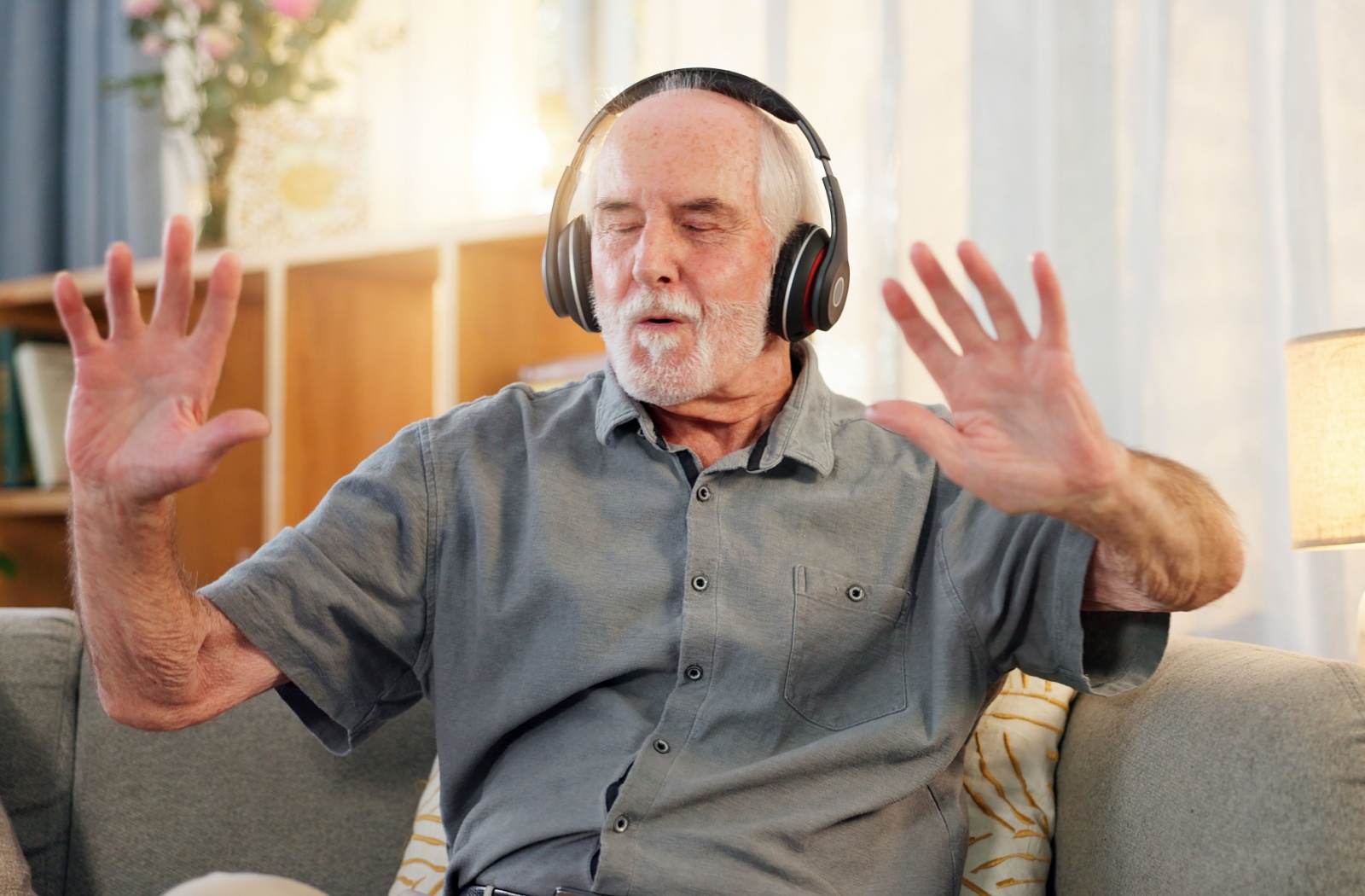 An older adult wearing black over-the-head headphones dances along to music while sitting on a living room couch