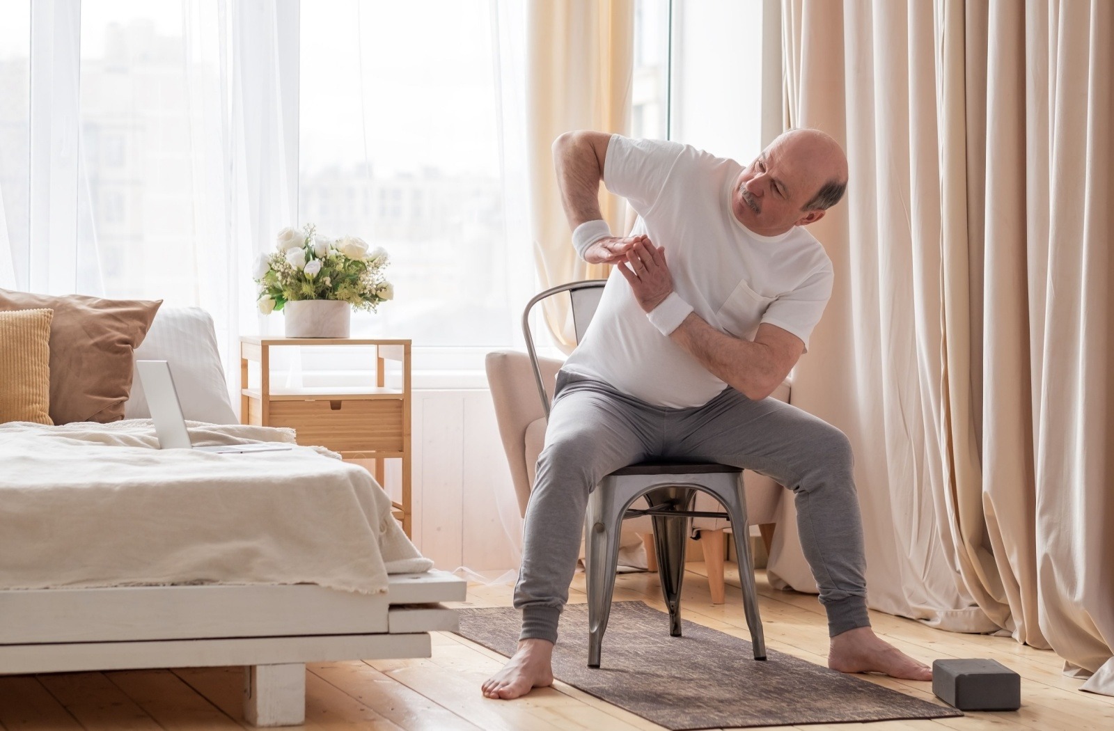 A senior performs a light stretch exercise while sitting on their chair in their bedroom.