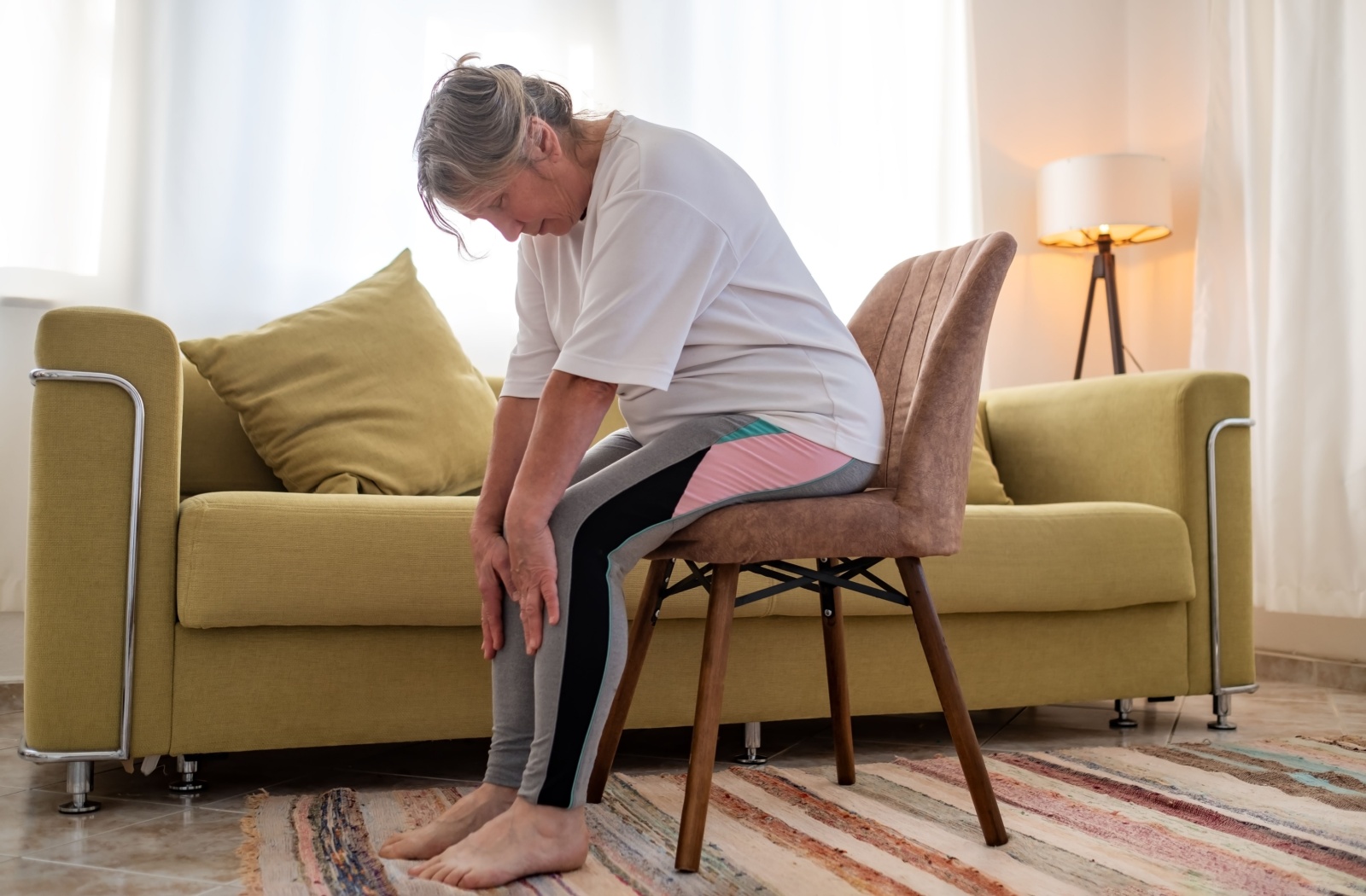 A senior in a white shirt sits on a chair in their living space and does a forward fold stretch.