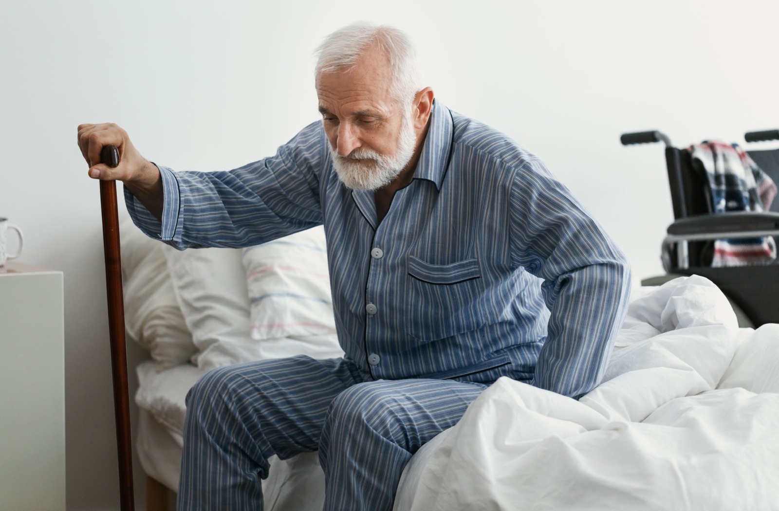 An older adult holding a walking cane in his right hand while sitting on a bed with a wheelchair beside it.