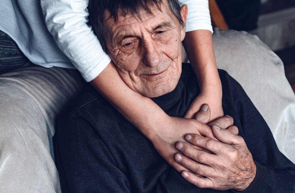 A family member putting their arms around an elderly parent sitting on a couch with their hands placed over each others.