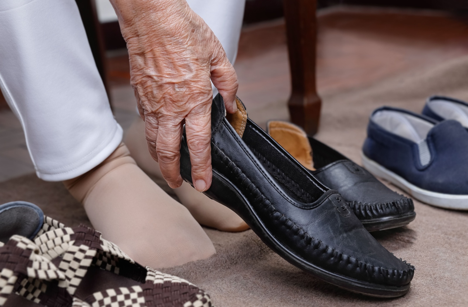 A close-up of an older adult bending over to put on a black slip-on shoe with their right hand.