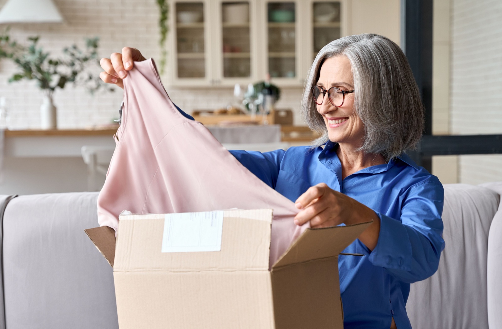 An older adult in senior living sitting on the couch and smiling as they open a box with a pink sleeveless shirt inside.