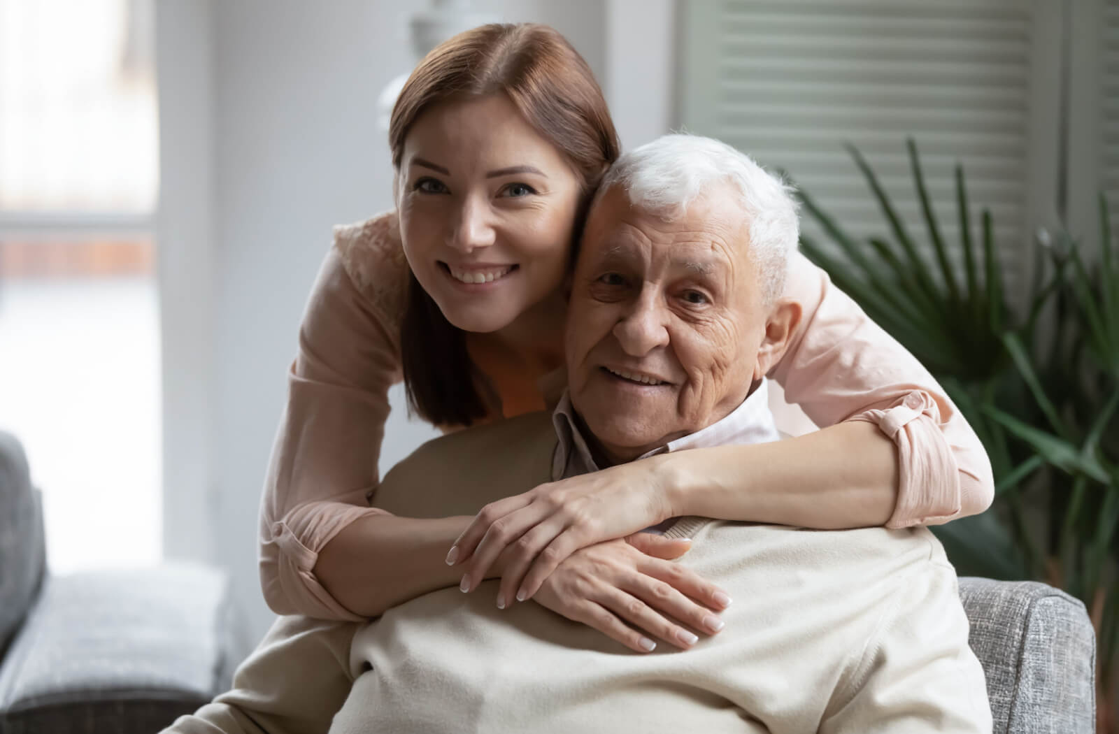 An older adult with memory loss smiling while their adult child hugs them from behind in a memory care community.