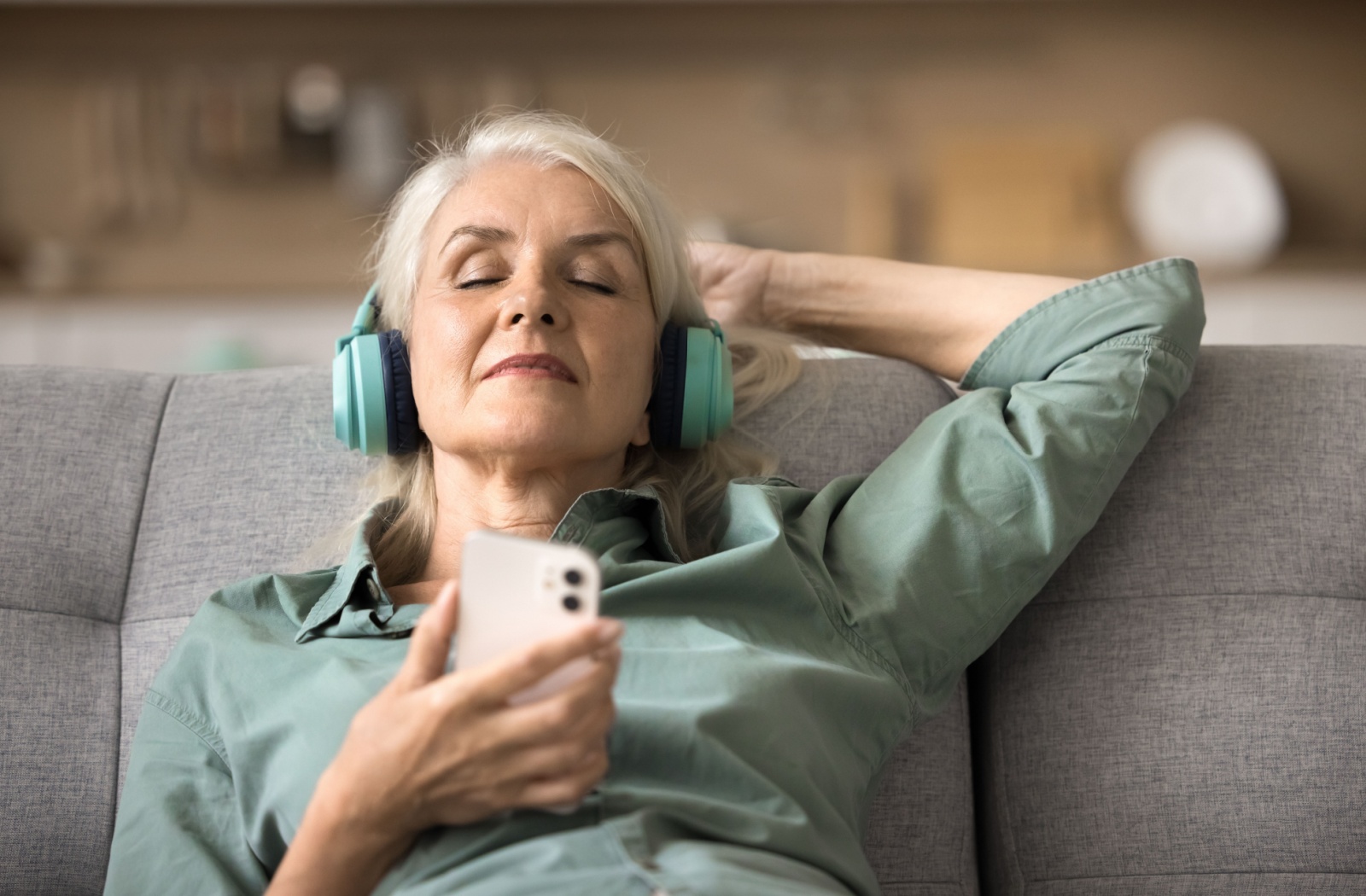 An older adult relaxing on their couch with headphones and their cell phone listening to an audiobook.
