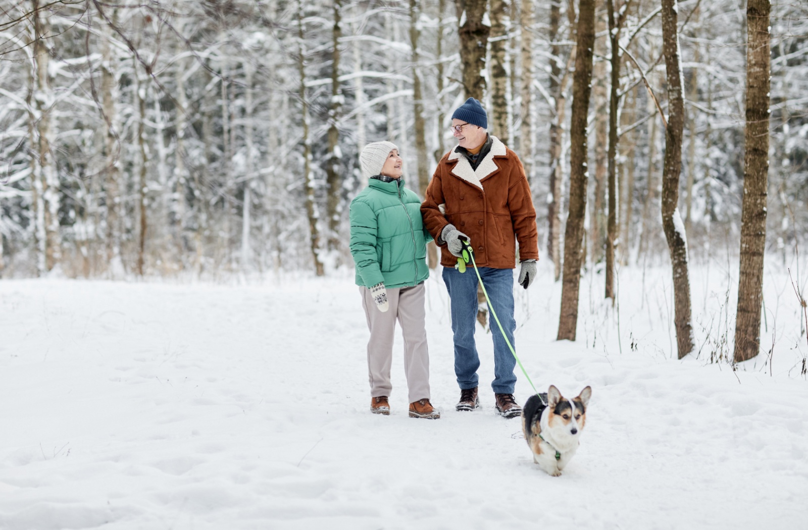 An older couple smile at one another while walking their small dog together through the snowy woods